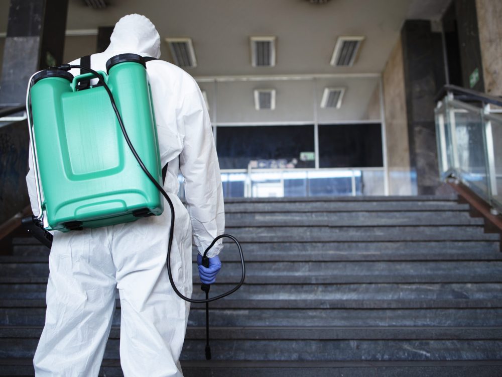 Unrecognizable person in white chemical protection suit with reservoir disinfecting public corridor to stop spreading highly contagious corona virus. Unrecognizable person in white chemical protection suit with reservoir disinfecting public corridor to stop spreading highly contagious corona virus.